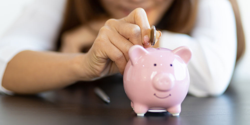 Nahaufnahme einer Frau, die eine Münze in ein rosafarbenes Sparschwein wirft. Der Fokus liegt auf der Hand und dem Sparschwein – das Bild steht symbolisch für Sparen und finanzielle Vorsorge.